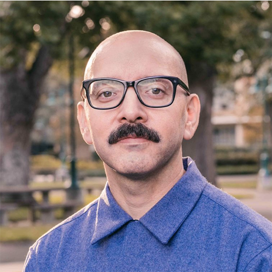 Headshot of a man with a bald head, black eyeglasses, and a thick moustache